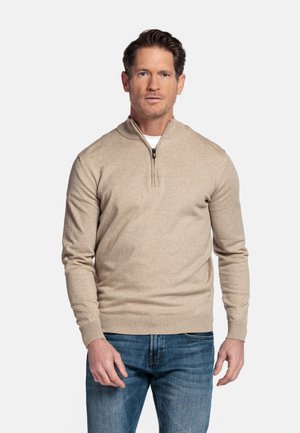 Man standing wearing beige quarter-zip sweater over white shirt and blue jeans, against plain light background.
