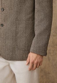 Brown herringbone jacket with three buttons, notched lapels, and a chest pocket, paired with light-colored trousers.