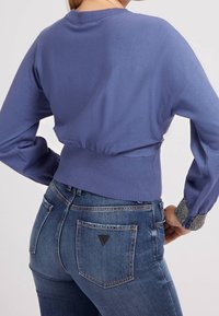 Woman wearing a fitted purple sweater with embellished cuffs and high-waisted blue denim jeans, shown from the back.