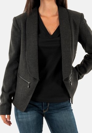 Mujer con blazer oscuro de cuadros con bolsillos con cremallera sobre una camiseta negra de cuello en V y vaqueros azules, de pie con una mano en la cadera.