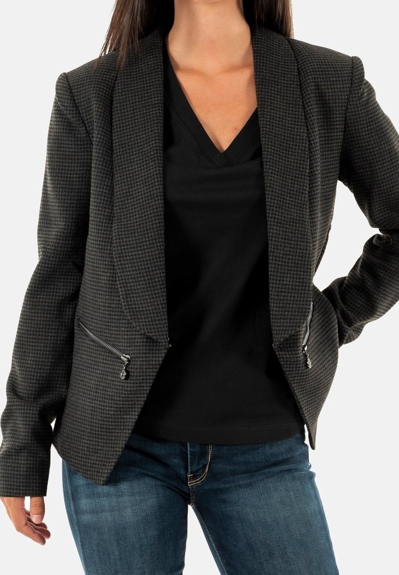 Femme portant un blazer à carreaux foncé avec des poches zippées sur un top noir à col en V et un jean bleu, debout avec une main sur la hanche.