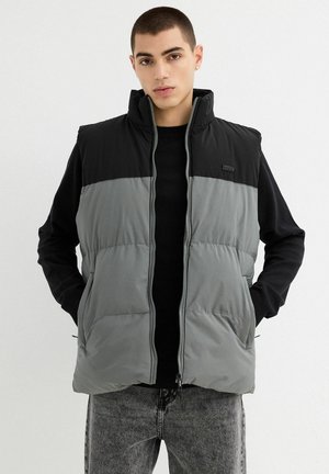 Chaleco acolchado gris y negro con cuello alto, cierre de cremallera, dos bolsillos laterales y un parche con el logo; llevado sobre un suéter negro.