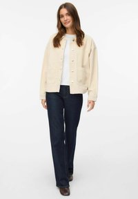 Beige kordøjjakke med frontknapper og sidelommer, parret med en hvid top og mørk denim jeans med lige ben.