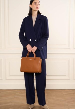 Blazer doppiopetto blu navy, top beige e pantaloni blu navy coordinati. Tiene una borsa in pelle marrone con due manici e dettagli del logo.