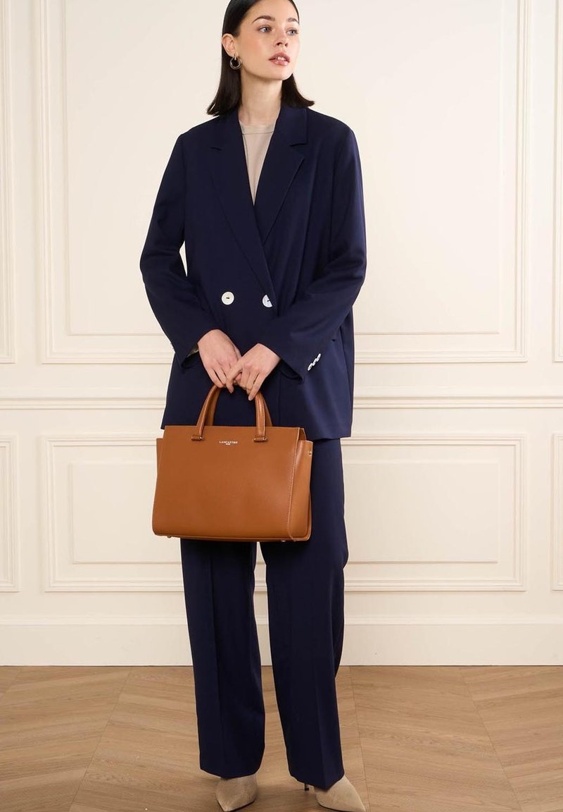 Blazer bleu marine à double boutonnage, haut beige et pantalon bleu marine assorti. Tient un sac à main en cuir marron avec des poignées doubles et un détail de logo.