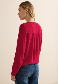 Chemisier rouge à manches longues avec un col rond, détail de pli au dos et tissu texturé. Assorti avec un jean en denim bleu.
