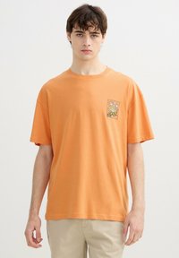 Joven con una camiseta naranja holgada con un pequeño gráfico de llama en el pecho, combinada con pantalones beige, de pie frente a un fondo liso.
