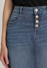 Falda de denim en azul lavado medio, que cuenta con una cintura alta, cuatro botones metálicos, una costura delantera y un bolsillo lateral. Textura suave y diseño ajustado.