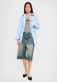 Žena v svetlomodrej oversized košeli cez pruhované tričko Moschino, s džínsovými kraťasmi po kolená a čiernymi špicatými balerínkami.