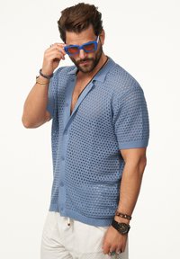 Hellblauer gestrickter Cardigan mit kurzen Ärmeln, offenen Mustern und Knöpfen. Kombiniert mit blauen Sonnenbrillen und weißen strukturierten Hosen.