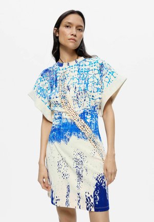 Femme portant une robe manches courtes avec des motifs abstraits bleus, blancs et beiges, debout devant un fond blanc.