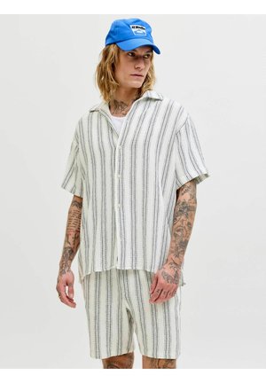 Persoon met tatoeages die een los wit overhemd met korte mouwen en korte broek met zwarte verticale strepen draagt, blauwe pet, kijkt opzij.