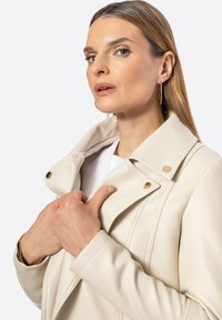 Krémová umělá kožená bunda s výstřihem, zlatými detaily a prošitím. Modelka drží límec oběma rukama.