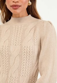 Beige Strickpullover mit hohem Hals. Verfügt über strukturierte Zopfmuster und gerippte Ärmel für zusätzlichen Detailreichtum und visuelles Interesse.