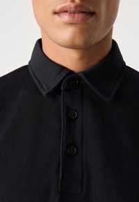Černé polo tričko se strukturovaným límcem, které má tři knoflíky a texturovanou látku. Detailní záběr zaměřený na límec a detaily knoflíků.