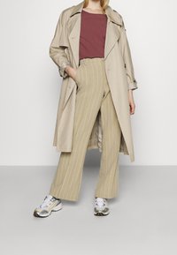 Beżowy płaszcz typu trench, bordowa bluzka i jasnobeżowe, szerokie spodnie w paski. Metaliczne srebrne sneakersy z żółtymi akcentami. Gładkie tekstury.
