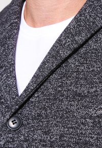 Primo piano di una persona che indossa un blazer testurizzato bianco e nero sopra una maglietta bianca a girocollo, evidenziando i dettagli dei bottoni e del risvolto.