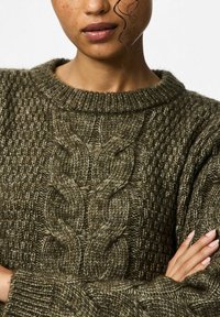 Mugav tumeoliiviroheline kudumiga sviiter ümmarguse kaelusega, millel on tekstuuriga keha ja silmapaistev nööride kudumismuster ees.