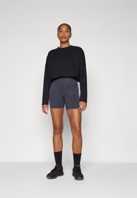 Zwarte lange mouwen cropped top gecombineerd met donkergrijze shorts. Model draagt zwarte sokken en schoenen, staande tegen een lichtgrijze achtergrond.