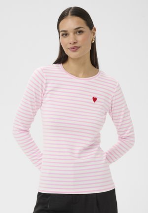 Ženska, oblečena v roza-belo črtasto majico z dolgimi rokavi in majhnim rdečim srcem na prsih, stoji z rokami za hrbtom.
