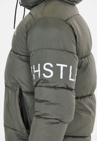 Olivgrön pufferjacka med quiltat mönster, vit "HSTL"-text på ärmen och en sidodragkedjeficka. Mjuk, isolerad tyg.