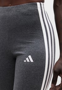 Κοντινή λήψη σκούρων γκρι αθλητικών κολαν με τρεις λευκές ρίγες στο πλάι και μικρό λογότυπο Adidas κοντά στο ισχίο, που φοριούνται από άτομο.