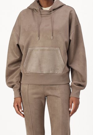 Osoba stojící před jednobarevným bílým pozadím, oblečená v taupe semišové mikině s kapucí a přední kapsou a odpovídajících kalhotách.