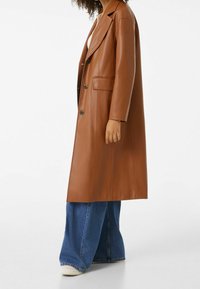 Hnědý dlouhý kabát z umělé kůže s klopami s výstřihem, předními kapsami a černými knoflíky, nošený přes volné modré džíny a bílé tenisky.