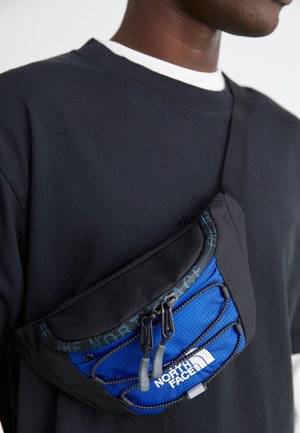 Henkilö, jolla on musta olkalaukku, jossa sininen etuosa ja valkoinen North Face -logo mustan paidan ja valkoisen kauluksen päällä.