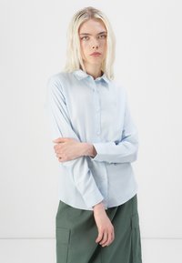Himmelblaues Hemd mit spitzem Kragen, langen Ärmeln und abgerundetem Saum, kombiniert mit grünen Cargo-Hosen. Glatte Stoffstruktur.