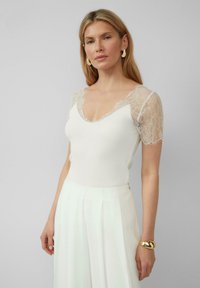Haut blanc côtelé avec des manches courtes en dentelle et un décolleté en V, associé à un pantalon blanc plissé taille haute. Bracelets dorés au poignet.