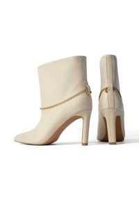 Valged nahast pahklaiused saapad terava ninaga, kõrgete stiletto kontsadega ja kuldse kettakujundusega pahkluu ümber, millel on kontrastsideme.