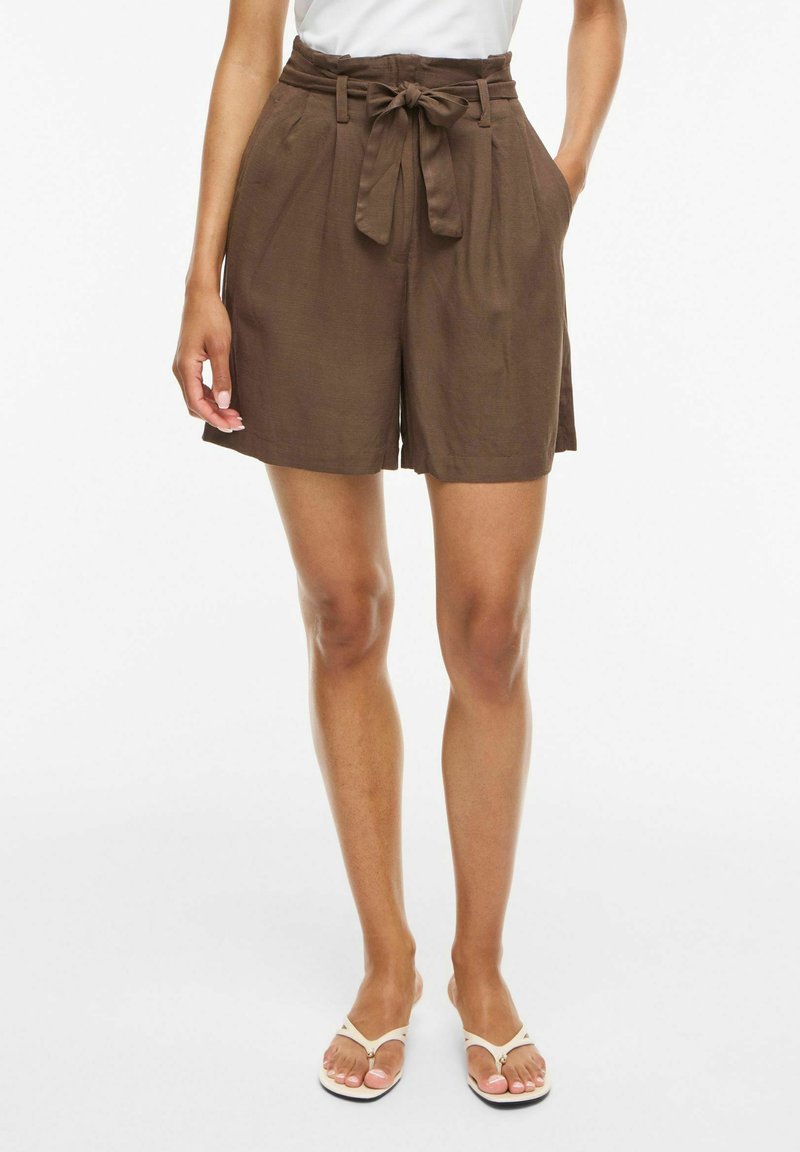 Femme portant un short marron taille haute avec ceinture à nouer devant et poches, associé à un haut blanc et des sandales blanches type tongs.