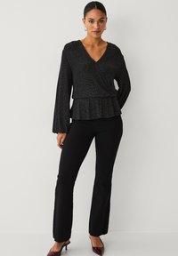 Schwarze glitzernde Wickelbluse mit langen Ärmeln und einem schwingenden Peplum-Detail, kombiniert mit eng anliegenden schwarzen Hosen und spitzen High Heels.