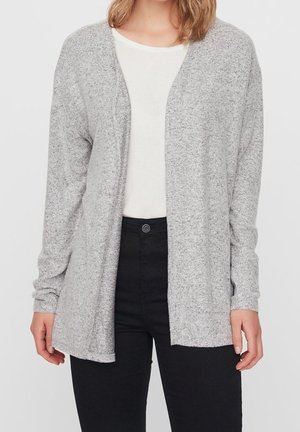 Kobieta ubrana w jasnoszary kardigan z otwartym przodem na białą koszulę i czarne spodnie z wysokim stanem na jednolitym tle.