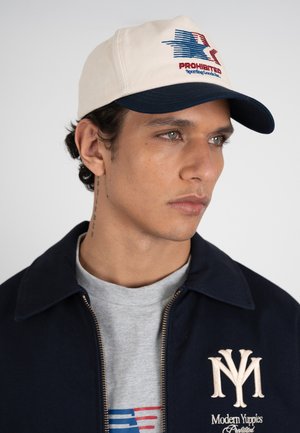 Nuori mies, jolla on beige ja laivastonsininen lippalakki, jossa on lipun logo, laivastonsininen takki, jossa on "Modern Yuppies" kirjailu, ja harmaa paita, jossa on punainen ja sininen kuvio.