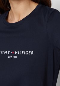 Námorně modré bavlněné tričko s kulatým výstřihem a vyšitým logem "TOMMY HILFIGER" v červených a bílých akcentcích. Založeno 1985.