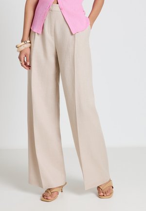 Donna che indossa pantaloni beige a gamba larga, top rosa senza maniche, sandali con tacco color cuoio e bracciali spessi al polso sinistro.