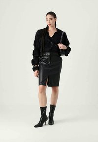 Γυναίκα με πλεξούδες, φορώντας ένα μαύρο αφράτο πουλόβερ, μαύρη δερμάτινη φούστα με ζώνη και μπροστινή σχισμή, μποτάκια και κρατώντας μια μικρή μαύρη τσάντα.