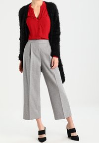Chemise rouge boutonnée, cardigan noir duveteux, pantalon large gris, chaussures noires à talons blocs ; tissu lisse avec des contrastes subtils de texture.