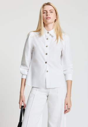 Blonde Frau trägt eine weiße, geknöpfte Bluse und weiße Hose, hält eine schwarze Handtasche vor einem schlichten Hintergrund.