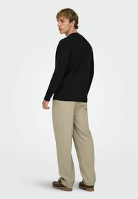 Man in beige pants, black sweater, and brown shoes standing, looking over left shoulder against a plain light background.
