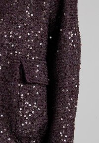 Tessuto in viola scuro decorato con paillettes, con superficie strutturata e una piccola tasca sul lato sinistro.