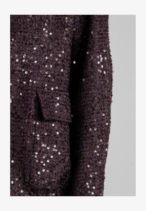 Tkanina zdobiona cekinami w ciemnym fiolecie, o fakturowanej powierzchni i małej kieszeni po lewej stronie.