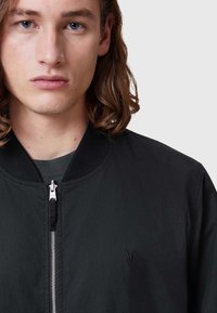 Schwarze Bomberjacke mit Reißverschluss, geripptem Kragen und gesticktem Akzent. Glattes Material mit matter Oberfläche, einfaches Design.