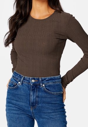 Mujer con un top marrón ajustado de manga larga con textura de ribete, metido dentro de unos vaqueros azul de tiro alto, con las manos apoyadas en las caderas.