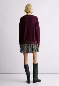 Maglione borgogna a maglia con una vestibilità ampia, sopra un vestito a balze stampato in bianco e nero, abbinato a stivali neri al ginocchio.