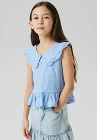Top senza maniche di colore azzurro chiaro con colletto a ruota e orlo a peplo. Il tessuto testurizzato presenta piccoli motivi floreali. Abbinato a una gonna di jeans.