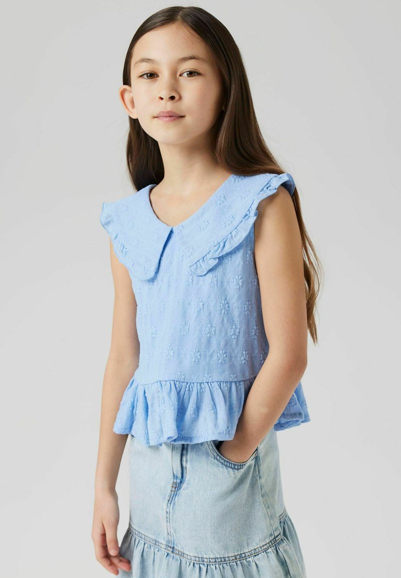 Top senza maniche di colore azzurro chiaro con colletto a ruota e orlo a peplo. Il tessuto testurizzato presenta piccoli motivi floreali. Abbinato a una gonna di jeans.