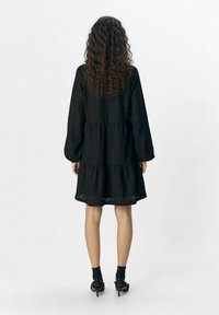 Robe noire à plusieurs niveaux avec des manches longues, dotée d'un tissu texturé. Portée avec des chaussettes noires jusqu'aux chevilles et des talons ouverts.
